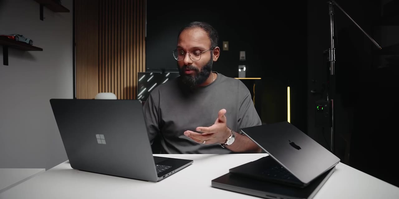 The presenter gestures beside two laptops on the desk: a Windows laptop on the left and a MacBook Pro on the right, setting up the side-by-side comparison.